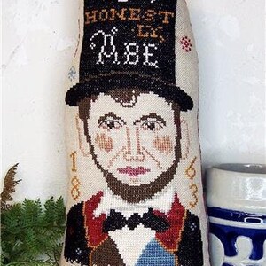 Puede incluir: Una almohada de punto de cruz con un retrato de Abraham Lincoln con un sombrero de copa. El texto en el sombrero dice "Oh Honest Abe" y el texto debajo del retrato dice "Freedom 1863".