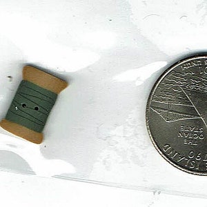 May include: A small, brown and green spool of thread with two holes in the side. The spool is sitting on a white surface next to a silver quarter with the text "Anne Frank House 2007" on it.