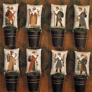 May include: Eight embroidered cushion ornaments featuring different depictions of Father Christmas. Each cushion is displayed in a small black metal bucket and surrounded by green pine branches.