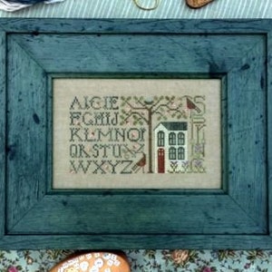 May include: A cross-stitch embroidery framed in teal blue wood. The design features a white house with a red door, a tree with birds, and the alphabet in a vintage font.