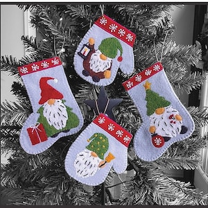 Peut inclure: Quatre décorations de Noël en feutre en forme de mitaines avec des visages de gnomes. Chaque gnome a un chapeau de couleur différente et un motif différent sur la mitaine. Les mitaines sont décorées de flocons de neige rouges et blancs.