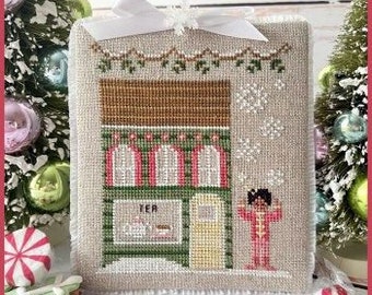 May include: A cross-stitch pattern featuring a small, green, and white building with a sign that reads "Tea & Cookies". The building has a red roof and a yellow door. A person in pink pajamas stands outside the building, blowing on their hands. The background is a white fabric with a snowflake and a garland of holly.