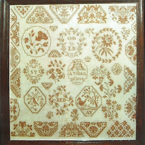 May include: A framed, antique sampler featuring various embroidered floral and geometric designs in shades of brown on an off-white background. The sampler includes text that reads "A token of love".