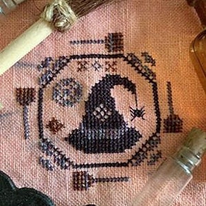 May include: A cross-stitch pattern featuring a witch's hat, broom, cauldron, and other witchy symbols. The design is stitched on a light pink fabric with brown and gray thread.
