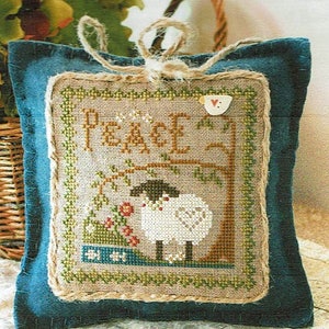 May include: A square, cross-stitched pillow with a teal blue border and a jute twine loop for hanging. The center features a sheep and tree design with the word "PEACE" stitched above. The design includes red and green floral accents.