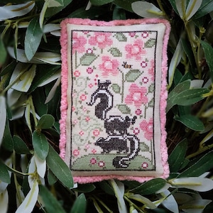 Può includere: Un cuscino ricamato a punto croce con un motivo floreale rosa e verde con tre puzzole nere e bianche. Il cuscino ha un bordo in velluto rosa.