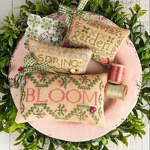 May include: A collection of embroidered decorative pillows with spring and garden themes. The pillows are beige with green and pink cross-stitch designs and text. A pink plate holds the pillows, surrounded by greenery and spools of thread.