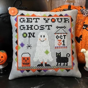 以下が含まれることがあります： 「GET YOUR GHOST ON」の文字が入った、ハロウィーンをテーマにした灰色の装飾用クッション。クロスステッチのゴースト、黒猫、ジャック・オー・ランタン、クモ、そして「OCT 31」と書かれたバナーが特徴です。