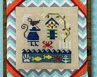 May include: Cross-stitch design featuring a whimsical beach scene with a cat, lighthouse, crab, and fish. The design is framed with a wooden border, orange ribbon, and a blue and white chevron pattern.