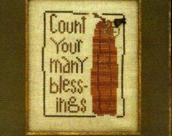 Op de afbeelding: Een ingelijst kruissteekkunstwerk met de woorden "Count Your Many Blessings" in bruin garen. Rechts van de tekst staat een bruin gestikt ontwerp. Het kunstwerk is ingelijst in een donkergroene lijst met een gouden binnenrand.
