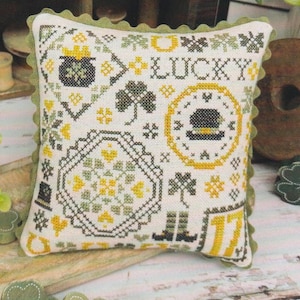 Puede incluir: Un cojín cuadrado de punto de cruz blanquecino con un borde festoneado verde. El diseño presenta un pote de oro, tréboles, un sombrero de duende, botas y la palabra "LUCKY" en negro y amarillo. También es visible el número "17".