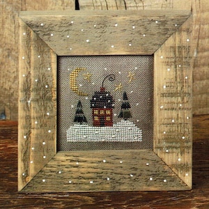 May include: A framed cross-stitch design featuring a small house with a chimney, two trees, a crescent moon, and stars against a snowy background.