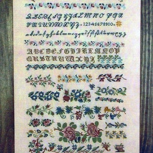 May include: A vintage sampler with a white background and a brown wooden frame. The sampler features a variety of embroidered designs, including letters, numbers, and floral motifs. The text on the sampler reads "ABCDEFGHIJKLMNO PQRSTUVWXYZ. 1841. 2. B.C.D.F.G.H.I.J.K.L.M.N.O.P.Q.R.S.T.U.V.W.X.Y.Z. 12345678910. a.b.c.d.e.f.g.h.i.j.k.l.m.n.o.p.q.r.s.s.t.u.v.w.x.y.z. ABCDEFGHIJKLMNOP QRSTUVWXYZ. J. Gebhardt."