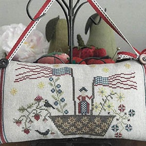 Op de afbeelding: Een handgestikt kussen met een patriottisch thema. Het kussen heeft een rood, wit en blauwe Amerikaanse vlag, een vrouw in een rode jurk en een boot met bloemen en vogels. Het kussen is versierd met sterren en heeft een knoopsluiting.