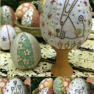 May include: A collection of handmade Easter eggs decorated with various embroidered designs. The eggs are made of fabric and feature a bunny, flowers, and a chick. The eggs are displayed on a wooden stand and a table.