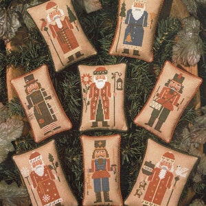 May include: Eight embroidered pillow ornaments featuring different Christmas characters. The ornaments are all rectangular and have a beige background with red, blue, and green accents. The characters include Father Christmas, a nutcracker, and a snowman.