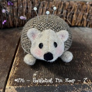 May include: A gray and brown hedgehog pin cushion with a black nose and white eyes. The pin cushion is 2 inches tall and has the text #676 - Hedgehog Pin Keep - 2" printed on the surface.