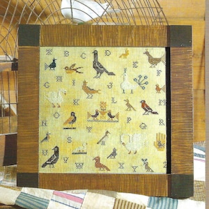 以下が含まれることがあります： 鳥や動物が描かれたアルファベットが刺繍された額装されたクロスステッチのサンプル。サンプルは薄い茶色の背景にクリーム色の生地でできています。文字は茶色のフォントで、鳥は赤、青、緑、黄など様々な色で描かれています。