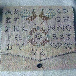 May include: A beige fabric needlepoint alphabet sampler with a floral design and two peacocks in the center. The sampler has a flap that folds over to create a small pouch.