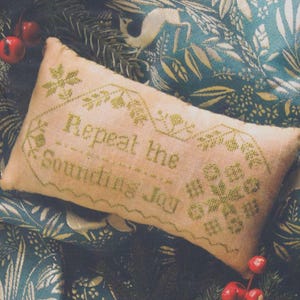 May include: A rectangular, light pink pillow with green cross-stitch embroidery. The text "Repeat the Sounding Joy" is centered, surrounded by floral and geometric designs. The pillow is set against a backdrop of teal fabric with a botanical pattern and Christmas decorations.
