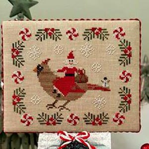 Op de afbeelding: Een kruissteekontwerp met een kardinaal die een kerstmanfiguur draagt, omringd door sneeuwvlokken, zuurstokken en hulst. Het ontwerp staat op een beige stof met een rode rand, perfect voor feestelijke decoratie.