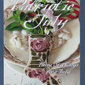 Puede incluir: Naturaleza muerta con un pequeño calcetín bordado con un diseño de rosas, lleno de flores y vegetación. El texto "Patriotic July" y "Three Stockings for July" son visibles. El fondo incluye un plato y una bandera americana.