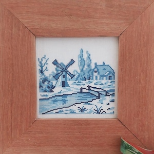 May include: A framed cross-stitch embroidery depicting a blue and white Dutch windmill and house with a river in the foreground. The embroidery is set against a white background and framed in a light brown wood frame.