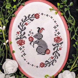 May include: Cross-stitch embroidery of a gray bunny holding a red flower, surrounded by a floral wreath with red roses and black accents. The embroidery is set in a pink oval hoop, perfect for home decor.