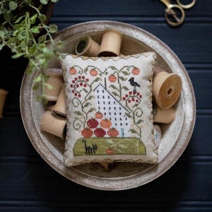 May include: A small, rectangular, cross-stitched pillow with a cream-colored background. The design features a house, berries, and a black bird. The pillow is surrounded by a scalloped edge and rests in a wooden bowl with spools of thread.