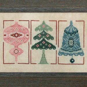 Puede incluir: Obra de arte de punto de cruz en un marco de madera. La obra presenta tres adornos navideños: una lágrima rosa, un árbol verde y una campana azul. Cada uno está enmarcado en un rectángulo rojo. El fondo es de tela beige claro.