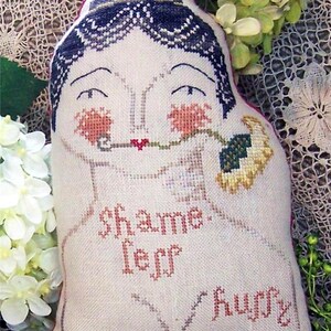 Puede incluir: Cojín decorativo con forma de rostro femenino, bordado a punto de cruz. El cojín tiene un fondo beige claro, cabello azul oscuro, mejillas rojas y las palabras "shame, less, hurry" bordadas en rojo. Con diseño floral.