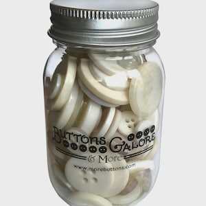 Peut inclure: Un bocal en verre transparent rempli de divers boutons blanc cassé. Le bocal a un couvercle en métal argenté et le texte "Buttons Galore & More" est imprimé sur le côté, ainsi qu'une adresse de site Web. Les boutons sont de formes et de tailles différentes.