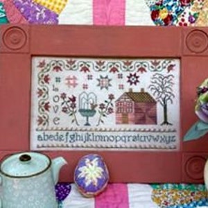May include: A framed cross-stitch embroidery featuring a house, a tree, a birdhouse, and a garden with flowers. The embroidery is in shades of pink, red, green, and blue. The frame is a reddish-brown color.