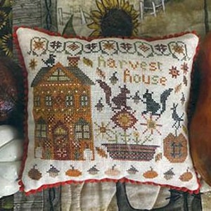 Puede incluir: Una almohada de punto de cruz con una casa marrón con chimenea, dos ardillas, una calabaza y el texto "Harvest House".