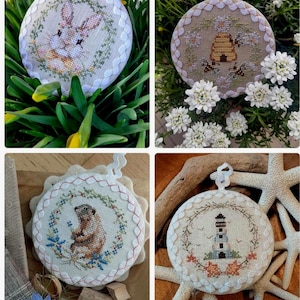 Może przedstawiać: Cztery okrągłe, haftowane ozdoby z białymi, falistymi krawędziami i pętelkami do zawieszania. Wzory obejmują królika, ul z pszczołami, świstaka i latarnię morską. Każda z nich otoczona jest kwiatowym wieńcem. Ozdoby umieszczone są na naturalnym tle.