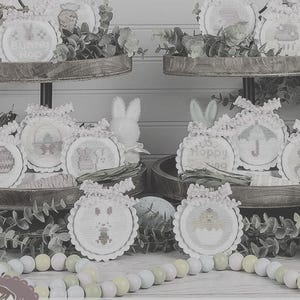 May include: A tiered display of Easter-themed cross-stitch ornaments. Each round ornament features a different design, including eggs, bunnies, and floral patterns. The ornaments are adorned with lace trim and ribbons, and are arranged with greenery and decorative beads.