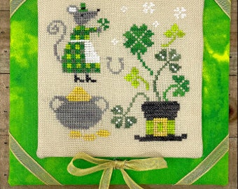 May include: A cross-stitch design featuring a green mouse holding a four-leaf clover, a pot of gold, and a shamrock plant in a leprechaun hat.