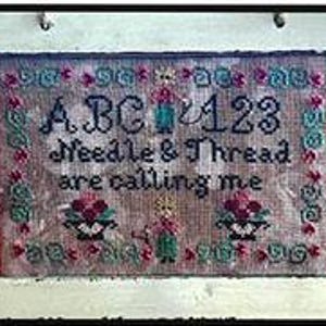 Op de afbeelding: Een ingelijst borduurwerk met de tekst "ABC & 123 Needle & Thread are calling me" en twee bloemmotieven. Het borduurwerk is in een wit frame met een rode en groene rand.