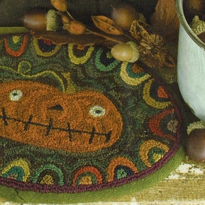 May include: Oval-shaped, hand-stitched pumpkin rug with a smiling jack-o'-lantern face. The rug features a green background with colorful, semi-circular patterns around the pumpkin. A decorative fall-themed item.