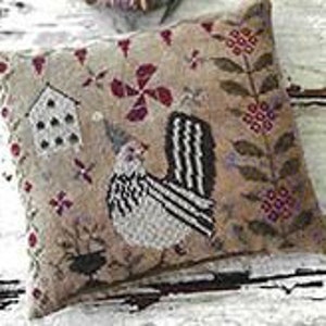 May include: A white and black rooster with a red party hat is embroidered on a beige fabric pillow. The pillow features a small house, flowers, and leaves in shades of brown, red, and green.