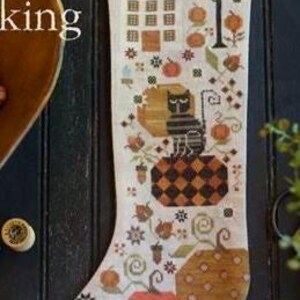 Könnte beinhalten: Ein dekorativer Weihnachtsstrumpf mit Herbstthema. Das Design zeigt ein Haus, Kürbisse, eine Katze und Krähen. Der Strumpf ist hauptsächlich cremefarben mit Akzenten in Orange, Braun und Schwarz. Der untere Teil des Strumpfes hat ein schwarz-weißes Zickzackmuster.