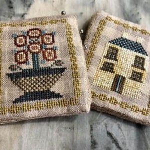 May include: Two small, square, beige fabric items with cross-stitch designs. One shows a floral arrangement in a basket, the other a house. Both have a gold border and are on a textured surface.
