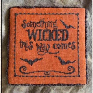 May include: Orange cross-stitch fabric with the phrase "Something Wicked This Way Comes" in dark gray. The design includes bats and decorative elements, with a gray border. A Halloween-themed decorative item.