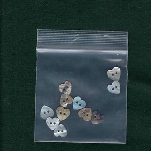May include: A plastic bag filled with a variety of heart-shaped buttons. The buttons are made of mother-of-pearl and have a iridescent sheen. Some of the buttons are a light blue color, while others are a brown color.