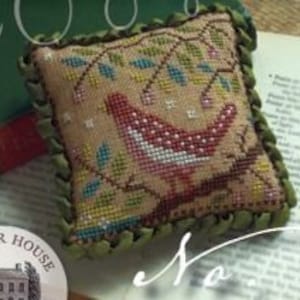May include: A cross-stitch pattern featuring a red bird with white spots perched on a branch with green leaves and pink flowers. The background is a beige fabric with a brown border.