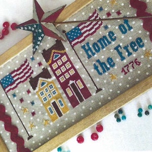 Puede incluir: Un diseño de punto de cruz enmarcado con una bandera estadounidense roja, blanca y azul, tres casas y el texto "Home of the Free 1776". El fondo es gris claro con puntos blancos.