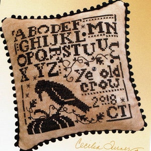 May include: A beige pillow with a black embroidered alphabet, a crow, and the text "Ye old crow" and "2018 CT". The pillow has a black pom-pom trim.