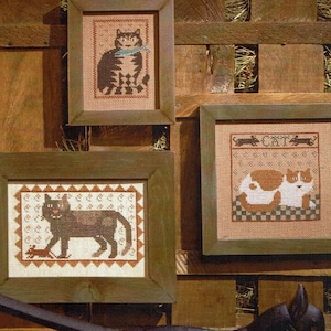 May include: Three framed cross-stitch cat pictures. One shows a brown tabby cat with a blue scarf. Another depicts a black cat with a mouse. The third shows two cats with the word "CAT" above them.