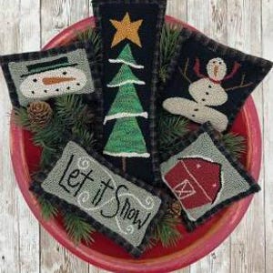Può includere: Una collezione di cuscini decorativi natalizi cuciti a mano, disposti in una ciotola rossa. I cuscini presentano disegni di un pupazzo di neve, un albero di Natale, un fienile e il testo "Let it Snow". La ciotola è circondata da rami di pino e una pigna.