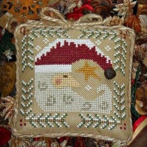 May include: Square, beige Christmas ornament with a cross-stitch design of Father Christmas. He wears a red hat with a white brim and holds a gold star. The ornament is framed with a green and white border and has a jute loop for hanging.